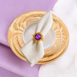 Rond de serviette doré avec pierre violette taillée, posé sur une serviette blanche sur une assiette dorée – décoration élégante de table pour mariage ou réception.
